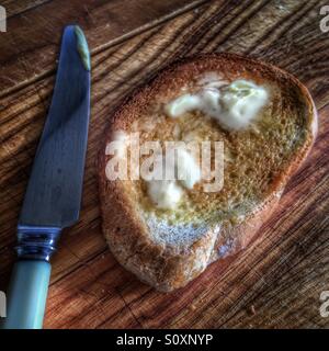 Pane tostato di mattina coperto con burro fuso visualizzato con coltello su una tavola di legno. Foto Stock