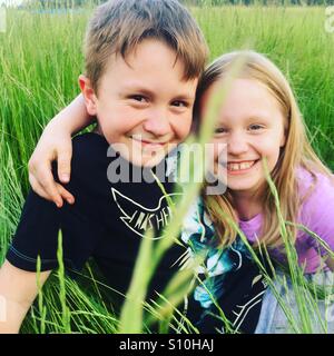 Fratelli di Felice costeggiata a vicenda e sorridente, seduti fuori in un alto erba verde su una multa, bello e soleggiato, fine settimana pomeriggio Foto Stock