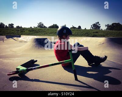 Un 9 anno vecchio ragazzo seduto da suo scooter a Eaton Park Skatepark in Norwich, Regno Unito Foto Stock