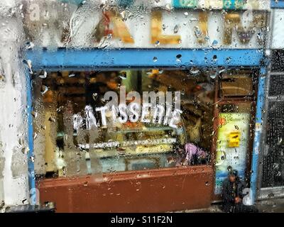 La donna al di fuori di un bagno turco bakers/pasticceria in un giorno di pioggia, visto attraverso un elevato wet finestra. , Harringay Green Lanes, London, Regno Unito Foto Stock