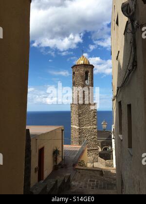 Guardando attraverso gli edifici per il campanile della cattedrale di Sant'Antonio Abate con il suo orologio e cupola a mosaico in Castelsardo, occidentale della Sardegna Foto Stock