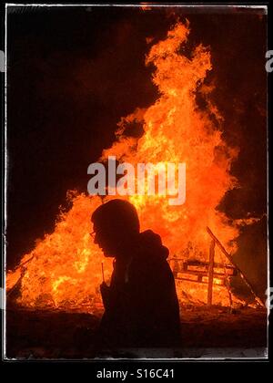 La silhouette di un uomo come lui cammina davanti a un falò infuria durante il 5 novembre celebrazioni (Guy Fawkes & notte dei falò). I pallet in legno vengono a bruciare in un incendio intenso. Credito foto © COLIN HOSKINS. Foto Stock