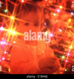 Una bambina davanti ad un luminoso albero di Natale con starbursts dalle luci Foto Stock