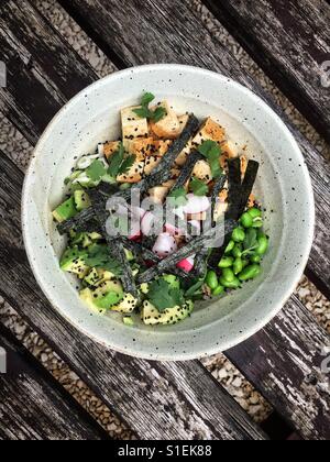 Un bianco ciotola di Hawaiian Poke con tofu, edamame fagioli, avocado, di semi di sesamo nero e strisce di nori alghe su un weathered tavola in legno rustico. Foto Stock