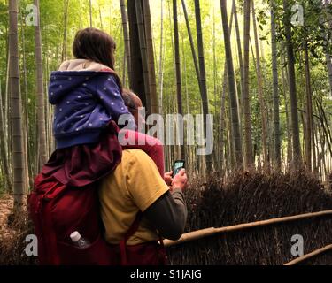 Bambina si siede sul suo padre le spalle mentre sta prendendo la foto con lo smartphone di foresta di bamboo a Kyoto, Giappone Foto Stock