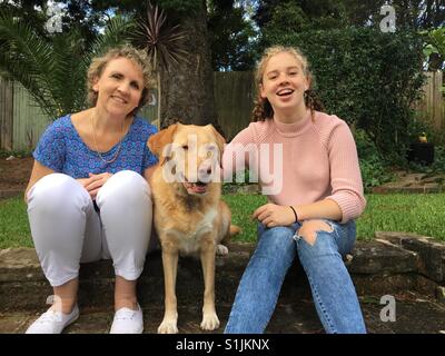 Australian madre e figlia adolescente seduto e sorridente con un cane in un cortile di casa Foto Stock