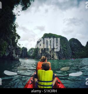 Una famiglia di un adulto e bambino in kayak sulle acque color smeraldo della baia di Halong in Vietnam. Picchi di calcare e rabboccato con la foresta pluviale. Foto Stock