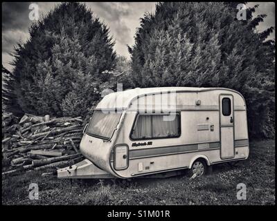 Un vecchio, abbandonato caravan si siede in un angolo di un campo, accanto a una pila di registri. Due grandi alberi sempreverdi forniscono la protezione. Credito foto - © COLIN HOSKINS. Foto Stock