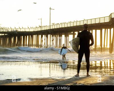 Padre orgoglioso permanente sulla spiaggia per guardare il figlio a piedi dalle onde dopo la navigazione. Tramonto a Pismo Beach pier, California USA Foto Stock