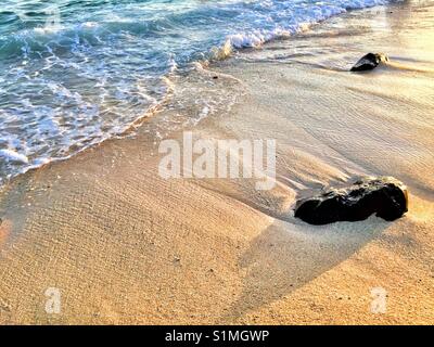 Pietre lavato fino a una spiaggia con surf a laminazione al tramonto . Hawaii Foto Stock