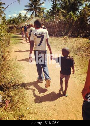 Uomo con una maglietta di Beckham a piedi con un giovane bambino attraverso un villaggio rurale in Chiure, Mozambico, Africa Foto Stock