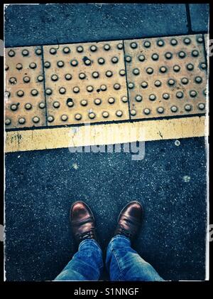 Un maschio di jeans blu e marrone e scarpe in pelle e in piedi su una piattaforma del treno con giallo con pavimentazioni tattili, London, Regno Unito Foto Stock