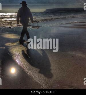 Silhouette e ombra di un uomo in un cappello a piedi lungo la spiaggia. Foto Stock