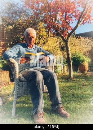 Uomo anziano seduto in giardino a leggere un libro, godendosi il sole autunnale Foto Stock