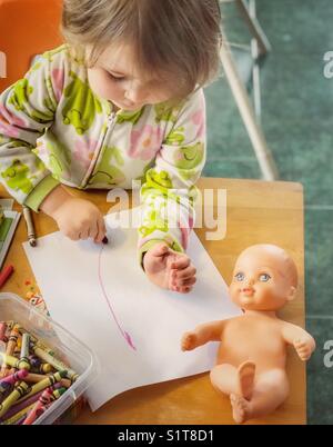 Il Toddler girl colorazione con una matita su un tavolo di legno con un baby doll su di esso Foto Stock