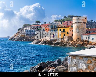 Perfetto piccolo villaggio sul mare di "Giglio Porto", con case multicolori, antica torre difensiva e una costa rocciosa contro un profondo blu del mare Mediterraneo. Isola del Giglio, Toscana, Italia Foto Stock