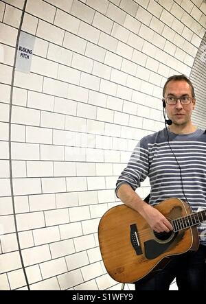 Un suonatore ambulante in un grigio e bianco striato pullover di eseguire con una chitarra acustica in mattonelle bianche nel tunnel della metropolitana di Londra ad un ufficiale di busker pitch Foto Stock