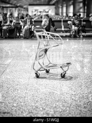 Svuota carrello bagagli nella sala partenze dell'aeroporto con righe di passeggeri seduti in background. Foto in bianco e nero Foto Stock