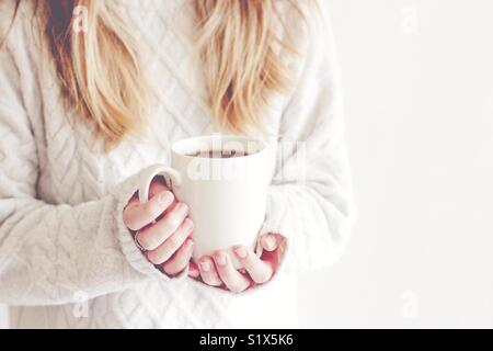 Bionda ragazza dai capelli in un accogliente Arran maglione tenendo una tazza di caffè caldo nelle sue mani in un tasto hi, immagine dello stile di vita Foto Stock