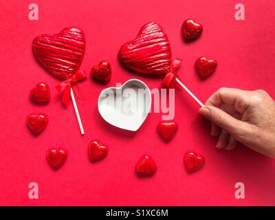 A forma di cuore, stagnola avvolto, cioccolato lecca-lecca, in una donna di mano con cuori di cioccolato, lecca-lecca e cuore in ceramica piatto di forma Foto Stock