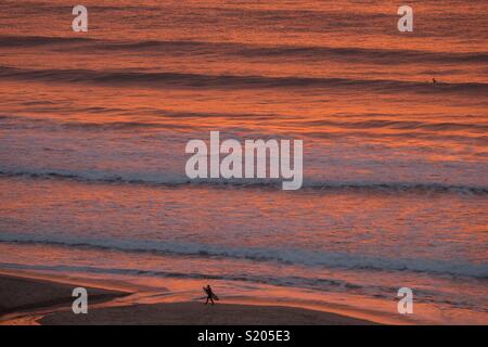 Un surfista si affaccia al mare come il sole tramonta sopra le onde a Sennen Cove, in Cornovaglia il 5 aprile 2018 in Inghilterra, Regno Unito. Foto Stock