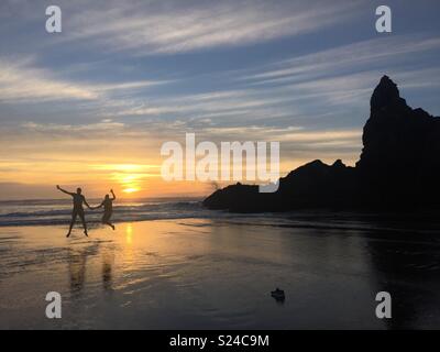 Due persone che saltano come il sole tramonta nel mare dietro di loro - Piha Beach, Auckland, Nuova Zelanda Foto Stock