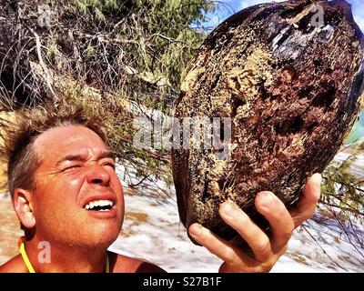 Conciate uomo inquisitively racchiude accolto intonacato di noce di cocco con sabbia su un deserto tropical island beach Foto Stock