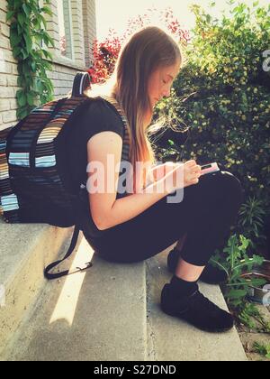 Tween schoolgirl con lunghi capelli biondi seduta sul passo indossando uno zaino guardando smartphone Foto Stock