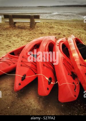 In plastica rosso kayaks ricreative impilati in fila su una spiaggia deserta a bassa marea in un giorno di pioggia. Spiaggia a mezzaluna, vicino a Vancouver, British Columbia, Canada. Foto Stock