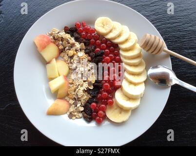 Vista in elevazione su piastra bianca con legno cucchiaio di miele, cereali e frutta selezionata per la prima colazione Foto Stock