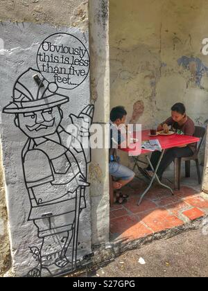 Un locale giovane avente il pranzo accanto a uno di George Town iconici di metallo sculture arte in Penang, Malaysia. Foto Stock