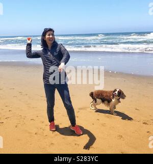 Giovane donna asiatica giocare a palla con English Springer Spaniel cucciolo di cane su una spiaggia di sabbia dorata sotto azzurro cielo blu in California Foto Stock