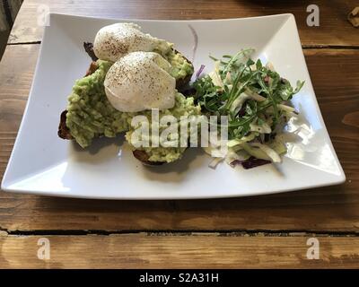 Pepati Uovo affogato con purea di avocado su pane tostato e un lato di insalata preparati tutti su una piastra bianca in appoggio su un tavolo di legno. Foto Stock