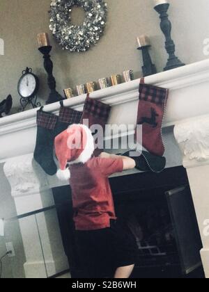 Little Boy vestito con un cappello da Babbo Natale che indossa una maglietta rossa e pantaloncini neri appendere le calze dal camino mantel per natale - Foto Stock