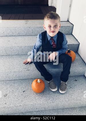 Ragazzino con capelli biondi, indossa una tuta blu e scarpe a scacchi seduto sui gradini della cappella in una Chiesa in America- seduto accanto a zucche su una bella caduta nitido pomeriggio Foto Stock