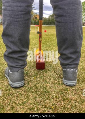 Tra le gambe vista posteriore di croquet gioco come giocatore colpisce la palla con un mazzuolo. Il Croquet è una strategia / sport giocato all'aperto che coinvolga colpire le sfere con un mazzuolo attraverso le cerchiature sul prato. Foto Stock