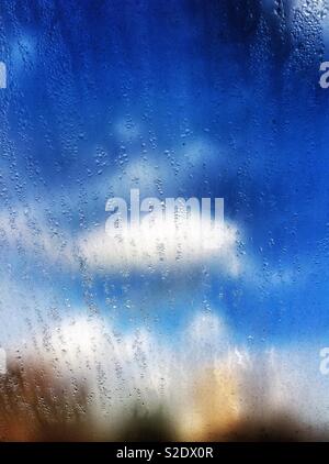 Nuvole bianche e blu cielo visto attraverso una sporca sporca finestra gocciolamento con umidità Foto Stock