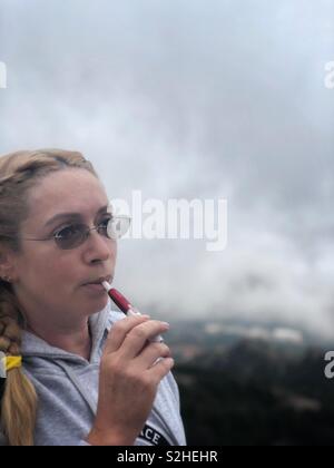 Giovane donna in bicchieri fuma sigaretta elettronica su un nuvoloso nebbioso giorno Foto Stock