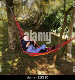 Bambino di nove anni utilizzando una compressa rilassante in un giardino amaca. Foto Stock