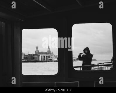 Vista degli edifici di fegato attraverso Mersey ferry finestra con uomo prendendo una foto in altra finestra. Lo skyline di Liverpool Foto Stock
