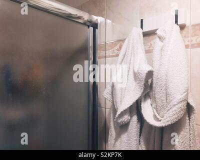 Un paio di accappatoi bianchi appesi al muro vicino al vetro della porta di un box doccia Foto Stock