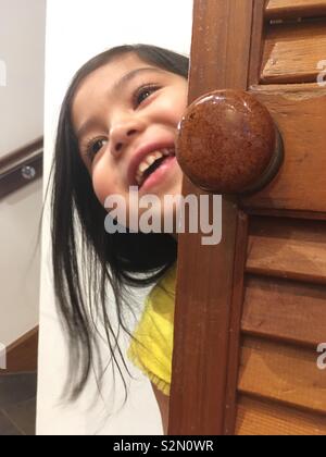 Un giovane multi-etnico (Caucasica/Punjabi/vietnamita) ragazza sorridente mentre il peering intorno a una cucina dispensa porta. Foto Stock