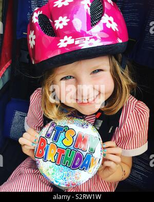 Bambina di cinque anni con un sorriso e indossando un sovradimensionamento / oversize Buon compleanno è il mio compleanno il badge / grandi badge mentre è seduto in una bicicletta rimorchio bici e indossa un casco ciclo / hat Foto Stock