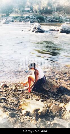 Una donna in un cappello, pantaloncini e scalzi, seduto tra rocce presso il fiume americano nel nord della California, rilassante Foto Stock