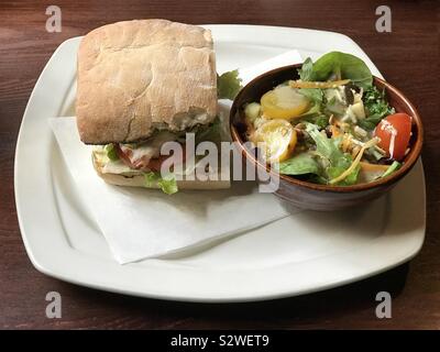Sandwich di Pollo su ciabatta pane e una insalata di contorno sono mostrati su una piastra isolata su un ristorante tavolo. Foto Stock