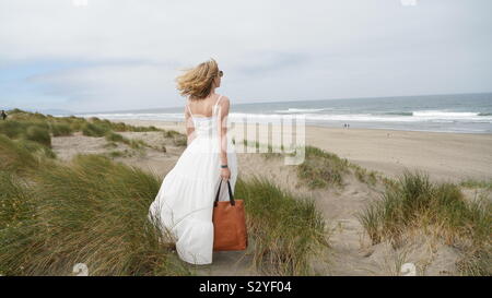 Giovane donna bionda, capelli soffiando nel vento, indossando lungo abito bianco, tenendo un marrone borsa in pelle, guardando a ocean da Sandy Hill Foto Stock