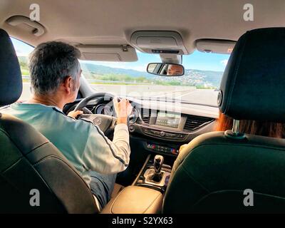 Un uomo alla guida di una auto a noleggio su una autostrada nel nord della Spagna in una giornata di sole con una luce calda come si vede dalla parte posteriore del sedile passeggero. Foto Stock