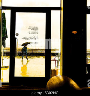 Visualizza la finestra di vetro portiera di un moderno cafe' di un pedone a piedi passando da sotto la pioggia con ombrello da mare lungomare Foto Stock