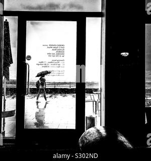 Visualizza la finestra di vetro portiera di un moderno cafe' di un pedone a piedi passando da sotto la pioggia con ombrello da mare lungomare Foto Stock