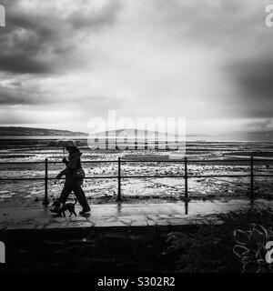 Un vivace passeggiata sul mare lungo la costa la passeggiata lungomare in wet Rainy day meteo tenendo un ombrello con un cane accanto al tallone Foto Stock